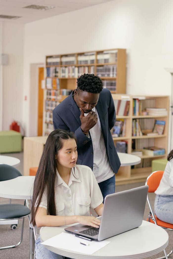 Teacher helping a student on a laptop in library classroom, fostering learning.